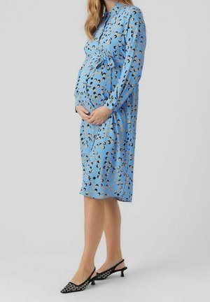 Kobieta w ciąży w niebieskiej sukience w wzory do kolan z długimi rękawami i czarnych niskich butach typu slingback, stojąca bokiem.