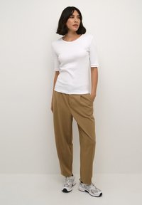 White fitted t-shirt with 3/4 sleeves, paired with loose beige trousers. Model wears gray sneakers with black accents. Plain background.
