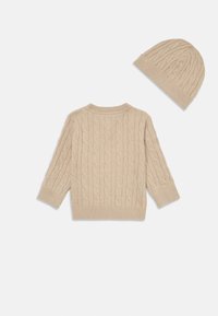 Un pull en tricot beige avec un motif en câble, des manches longues et des poignets côtelés, associé à un bonnet assorti. Les deux articles présentent une texture douce.