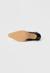 Zapato de tacón alto negro con punta afilada y parte superior de cuero liso. Presenta una suela color beige con ranuras y un acento de logo plateado.