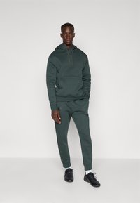 Mann stående mot en hvit bakgrunn iført en mørk grønn hettejakke og matchende joggebukser med svarte joggesko og hvite sokker.