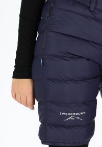 Pantaloni corti imbottiti blu navy in materiale sintetico, dotati di una tasca con zip, logo e un accento con la bandiera svedese vicino alla zipper.