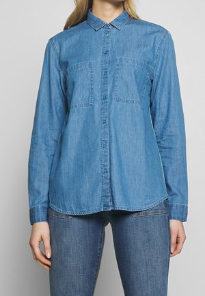 Kobieta ubrana w długi rękaw, zapinaną na guziki, niebieską dżinsową koszulę z dwoma kieszeniami na piersi oraz niebieskie dżinsy na jednolitym tle.