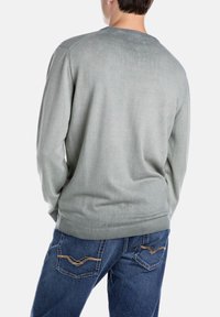 Helles graues Rundhals-Pullover mit glatter Textur. Die Rückansicht zeigt dezente Nähte, kombiniert mit blauen Jeans, die dekorative Taschen haben.