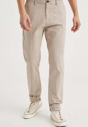 Beige, schlanke Hose mit dezenten vertikalen Streifen, umgeschlagenen Säumen, getragen mit cremefarbenen High-Top-Sneakern an einer stehenden Person.