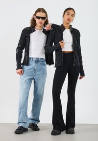 Sort ribbet zip-jakke med subtilt mønster, båret over en hvid crop top. Blå baggy jeans og sorte flarebukser. Kraftige sorte sko.