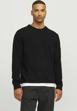 Schwarzer Pullover, gestrickte Textur, runder Halsausschnitt, lange Ärmel, schmal zulaufende Bündchen, über einem weißen Hemd getragen, teilweiser Blick auf graue Hose.