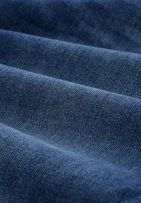 Džinsu audums dziļi zilā krāsā, ar teksturētu austu struktūru un smalkām variācijām, izlikts krokās, kas izceļ tā izturību un mīkstumu.