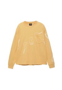 Chemise beige à manches longues avec une poche poitrine et des lettres blanches disposées en diagonale sur le devant et les manches.