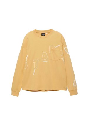 Beige pitkähihainen paita, jossa on rintatasku ja valkoisia kirjaimia vinosti ripoteltuna paidan etuosassa ja hihoissa.