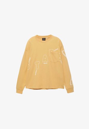 Beige pitkähihainen paita, jossa on rintatasku ja valkoisia kirjaimia vinosti ripoteltuna paidan etuosassa ja hihoissa.