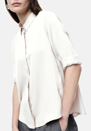 Donna che indossa una camicia bianca a maniche lunghe con bottoni e maniche arrotolate, pantaloni grigi, in piedi con una mano in tasca.