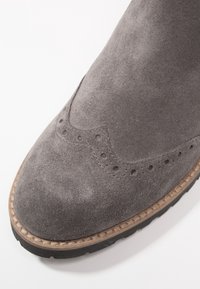 Grå mocka ankelsko med brogue-detaljer, rund tå och en kontrasterande ljusbrun sydd rand. Gummisula för bra grepp.