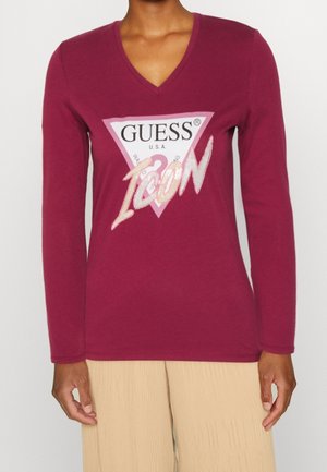 Femme portant un haut bordeaux à manches longues et col en V avec un logo Guess rose et blanc et le mot "ICON" imprimé à l'avant.
