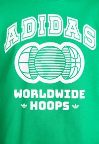 Žalias medvilninis marškinėliai su baltais užrašais "ADIDAS WORLDWIDE HOOPS" ir trimis stilizuotomis krepšinio grafikos. Audinys su banguota tekstūra.