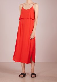 Robe midi rouge sans manches avec un design à volants superposés et une fente latérale, en tissu léger. Portée avec des sandales plates noires à enfiler.