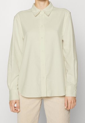 Osoba ubrana w długorękawową, jasnobeżową koszulę na guziki z kołnierzykiem, połączoną z kremowymi spodniami na jednolitym tle.