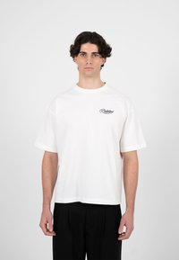 Ung mann med krøllete mørkt hår iført en hvit oversized T-skjorte med liten tekst på brystet og svarte bukser, stående mot hvit bakgrunn.