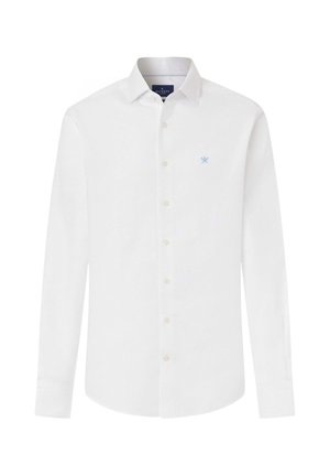 Joven con camisa blanca abotonada metida en pantalones azul marino, con zapatos marrones de vestir, de pie frente a un fondo claro y liso.
