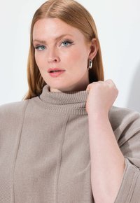 Jasnobeżowy sweter dzianinowy z wysokim, ribbowanym kołnierzem i teksturowanymi panelami. Model trzyma kołnierz jedną ręką, ukazując subtelne detale.