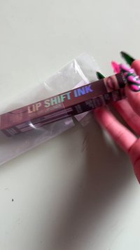 Een hand met lange, kleurrijke nagels houdt een lippenbalsemtube met het etiket "LIP SHIFT INK", die gedeeltelijk uit de verpakking is verwijderd, op een vlakke ondergrond.