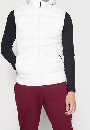 Mann in einer weißen gepolsterten Weste über einem schwarzen Langarmshirt und kastanienbraunen Hosen mit Text an der Seite, stehend mit einer Hand in der Tasche.
