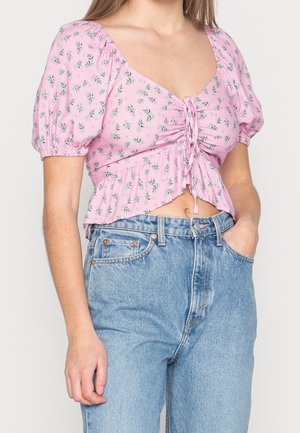 Mujer con un top corto floral rosa con mangas abullonadas y jeans de talle alto azul claro, con una mano en el bolsillo.