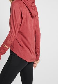 Red long-sleeve athletic hoodie with a textured finish, featuring a hood, side slits, and gold accent stripes on the sleeves. Paired with black leggings.