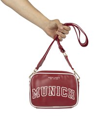 Borsa a tracolla in finta pelle rossa con rifiniture bianche, hardware dorato e "MUNICH SPORTS" stampato in lettere bianche in grassetto sulla parte anteriore.
