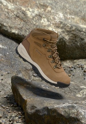Bota de senderismo marrón con suela negra y cordones estampados, situada sobre terreno rocoso, enfatizando su uso resistente al aire libre.