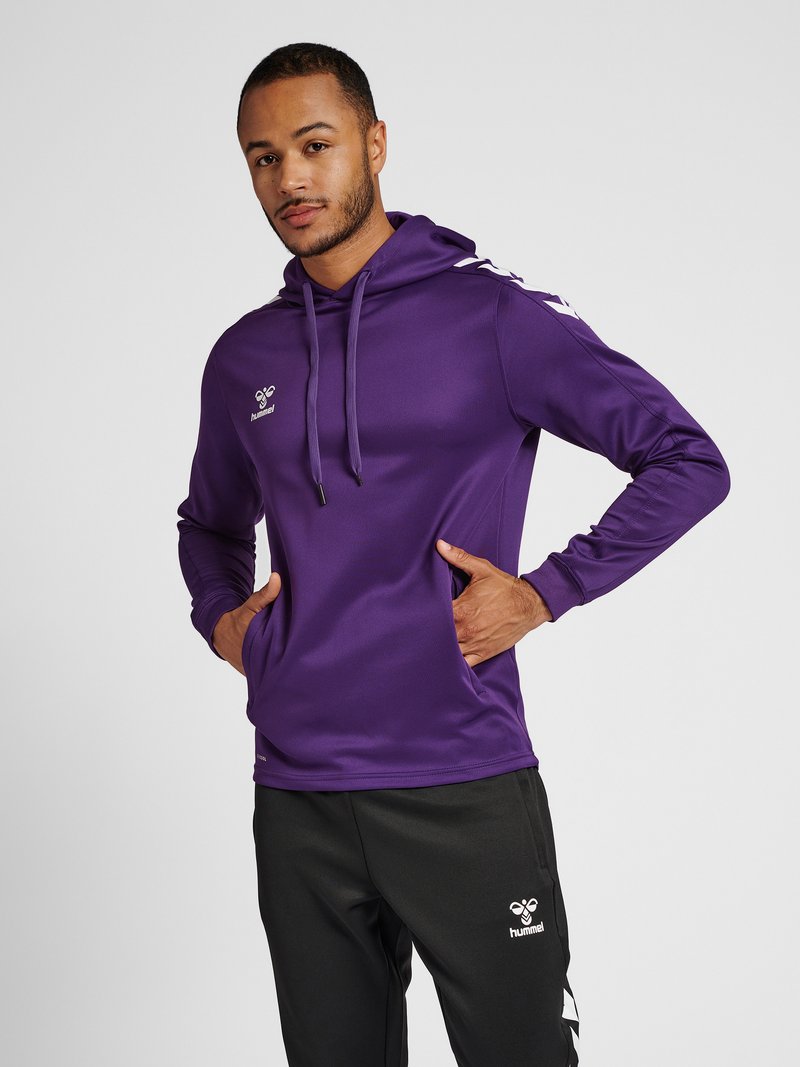 Mann trägt lila Kapuzensweatshirt und schwarze Hose mit weißen Markenlogos, steht mit den Händen in den Taschen des Sweatshirts vor weißem Hintergrund.