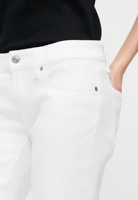 Μέση και πάνω μέρος μηρού ατόμου που φορά εφαρμοστά άσπρα τζιν και μαύρο τοπ, με το δεξί χέρι στην μπροστινή τσέπη.