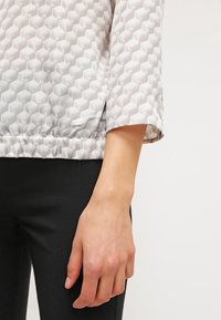 Haut à manches longues, de couleur gris clair, avec un motif hexagonal texturé, doté d'une taille élastique et d'une coupe ample, associé à un pantalon noir.
