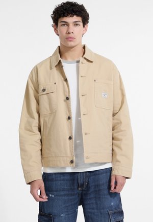 Giacca in canvas beige con chiusura a bottone, due tasche anteriori e una piccola etichetta sul petto. Indossata sopra una camicia grigia e pantaloni cargo blu.
