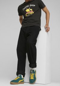 Black graphic t-shirt featuring a "Coffee Shop" design, paired with cuffed black jeans and green suede sneakers with yellow accents.