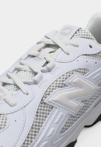 Κοντινό πλάνο ενός λευκού αθλητικού παπουτσιού με υφασμάτινο δίχτυ, επενδυμένη γλώσσα, κορδόνια και ανάγλυφο λογότυπο "N" στο πλάι.