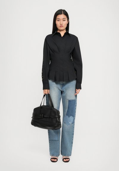 Femeie îmbrăcată cu o bluză neagră cu mâneci lungi și nasturi, blugi patchwork cu design în formă de inimă, tocuri negre cu vârf deschis, ținând o geantă mare neagră.