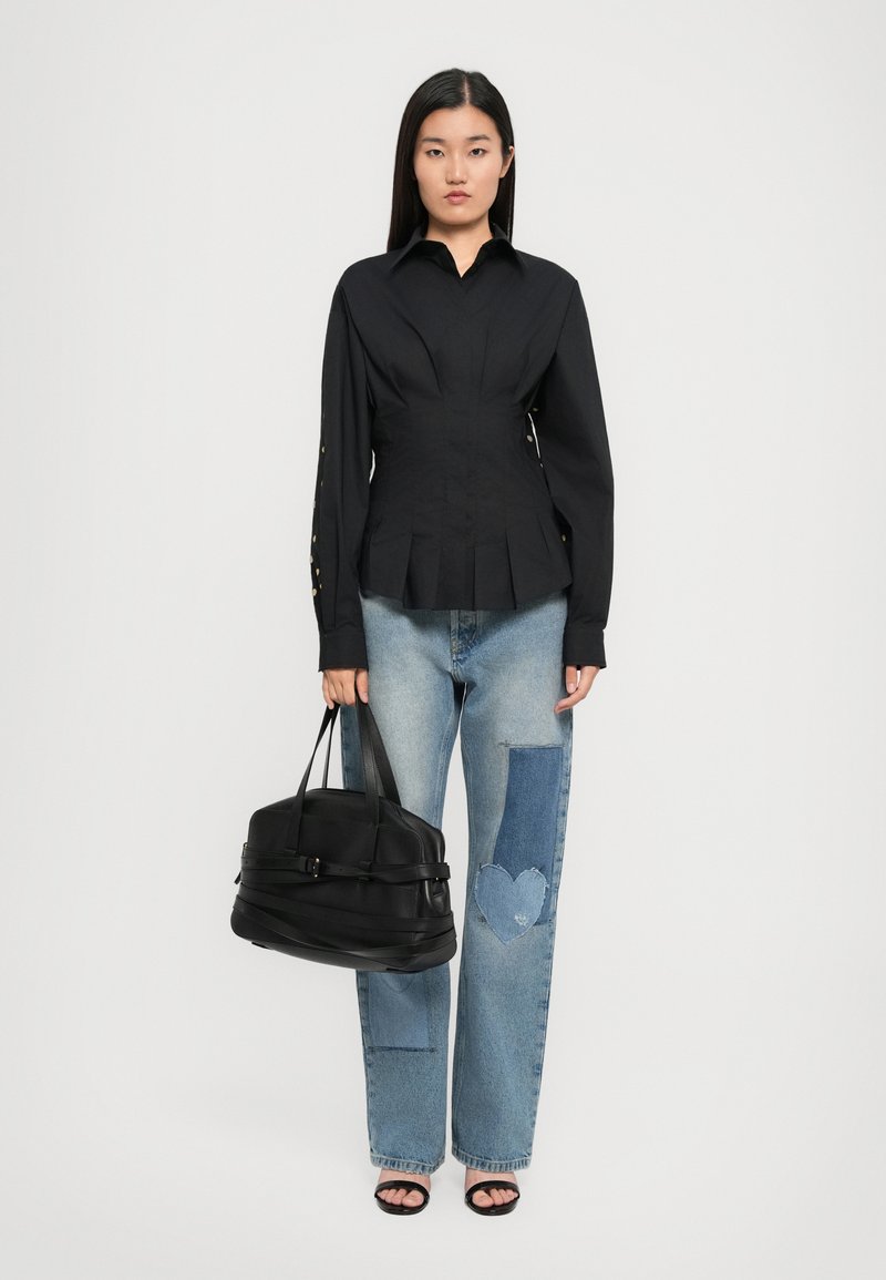 Vrouw in zwarte lange mouwen blouse met knopen, patchwork jeans met hartontwerp, zwarte open teen hakken, houdende een grote zwarte handtas.