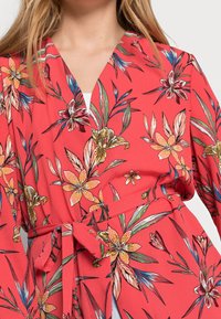Blomstret kimono i koralfarvet stof med bånd i taljen. Har et levende blomsterprint med gule, pink og grønne accenter. Blød, let tekstur.