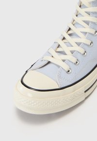 Světle modré plátěné tenisky s bílými tkaničkami, krémovým gumovým špičkou a texturovanou podrážkou. Má černé gumové detaily kolem okraje.