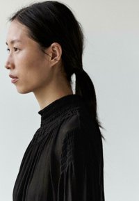 Sort plissert bluse med høj hals, lavet af tekstureret stof og lange ærmer, båret af en person med håret sat op, vendt sidelæns.