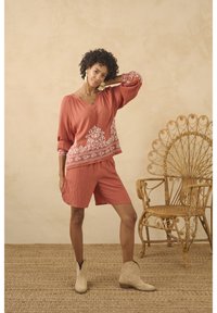 Korallenfarbener Oberteil mit weißer Blumenstickerei und voluminösen Ärmeln, kombiniert mit passenden plissierten Shorts. Beigefarbene Knöchelstiefel und ein Rattanstuhl im Hintergrund.