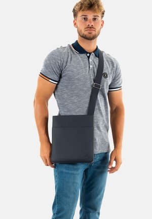 Borsa a tracolla in pelle blu scuro con una superficie liscia, forma rettangolare e tracolla regolabile a righe. Presenta un logo e una tasca frontale.