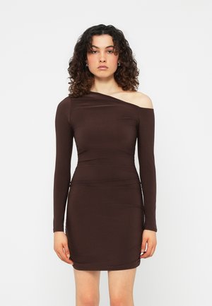 Mujer con cabello rizado que lleva un vestido mini ajustado de manga larga y color marrón oscuro, con un hombro descubierto, posando frente a un fondo blanco liso.