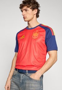 Röd och blå kortärmad tröja med gula accenter, med texturerad finish, gyllene emblem och vertikalt randmönster.