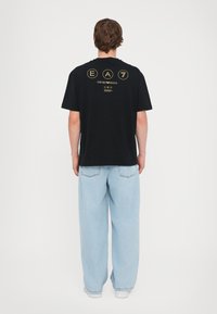 Black cotton t-shirt with short sleeves, featuring gold text and logos on the back. Worn with light blue wide-leg jeans.