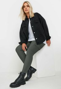 Svart denimöverdimensionerad jacka med silverknappar, matchad med olivgröna ribbade leggings och svarta snörstövlar på en vit block.
