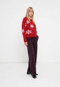 Pull rouge avec des motifs de flocons de neige blancs, associé à un pantalon large violet foncé et des chaussures noires à bout carré. Texture tricot lisse.