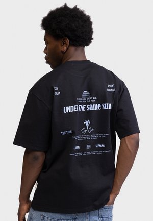 Ung mand med kort, krøllet hår iført en sort t-shirt med hvid tekst og palmetræ-design på ryggen.