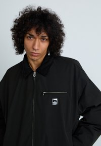 Casaco preto com fecho éclair e colarinho de veludo, apresentando um bolso no peito com um fecho prateado e um patch com o logótipo "OBEY". Textura suave.